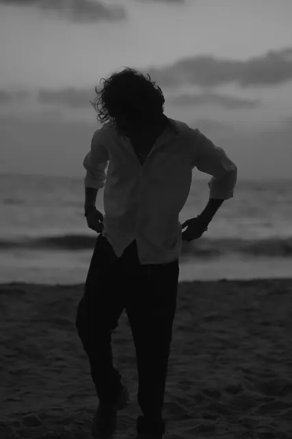 Black and white frame of a person standing on a beach at dusk, hands on hips, sea and low cloud behind them.