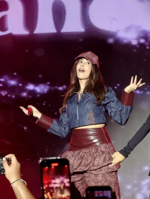 A performer in a cap and denim jacket mid-song on a bright stage, arms wide, phones held up in the crowd below.